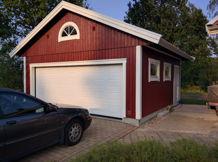 Nybyggt garage med röda väggar, vit garageport och vita fönster, beläget på en gjuten platta.