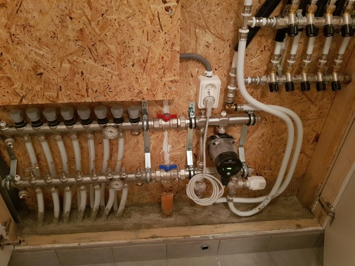 Golvvärmesystem med synliga rör och fördelare mot en OSB-vägg, förberett för läckagehantering.