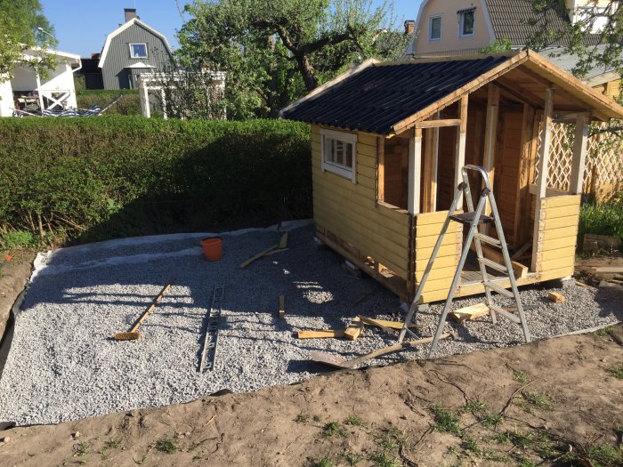 Gul stuga under konstruktion, grusgrund, trädgårdsarbete, stege, soldagsljus, häck, bostadshus i bakgrunden.