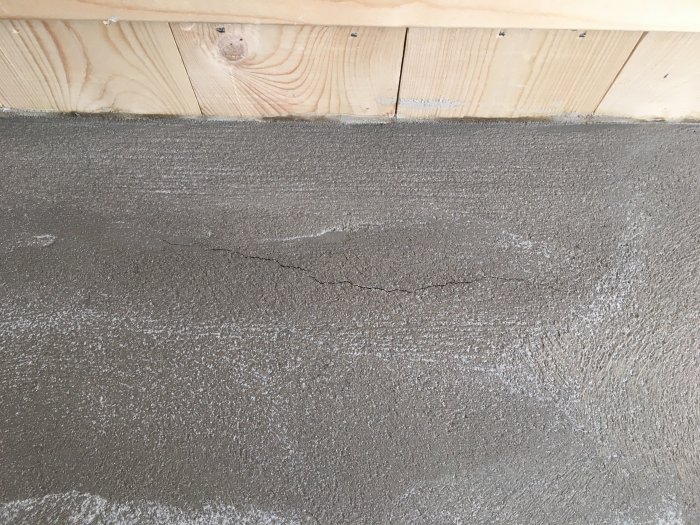 Närbild av grå putsad vägg med synlig sprickbildning och randmönstrad yta under träbjälke.