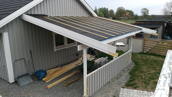 Nästan färdigbyggd carport med monterad takstol och elcentral, bredvid ett grått hus.