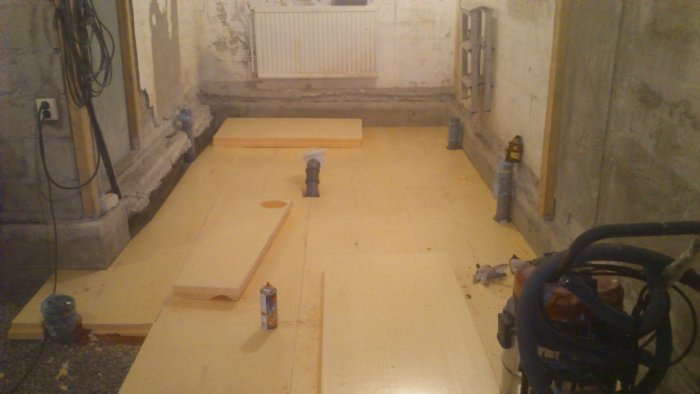 Källarrenovering med gul cellplast på golvet, rörstump och byggmaterial synliga.