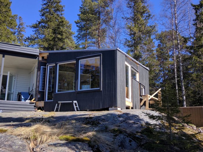 Ett modernt svart trähus med stora fönster omgivet av träd under en klarblå himmel.