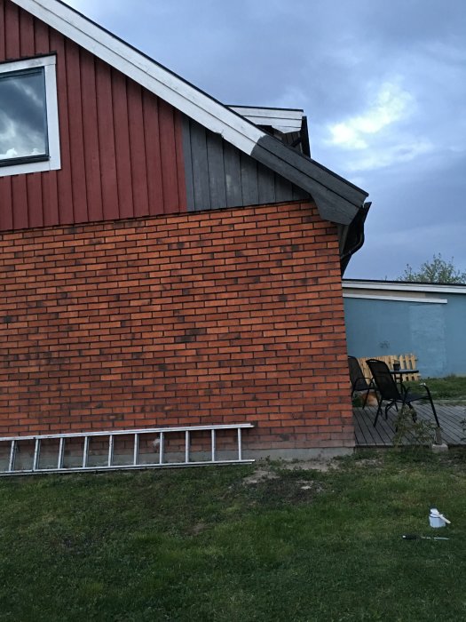 Hörn av ett hus med tegelvägg och del av fasad med provmålad yta, stegar och målartillbehör på marken.