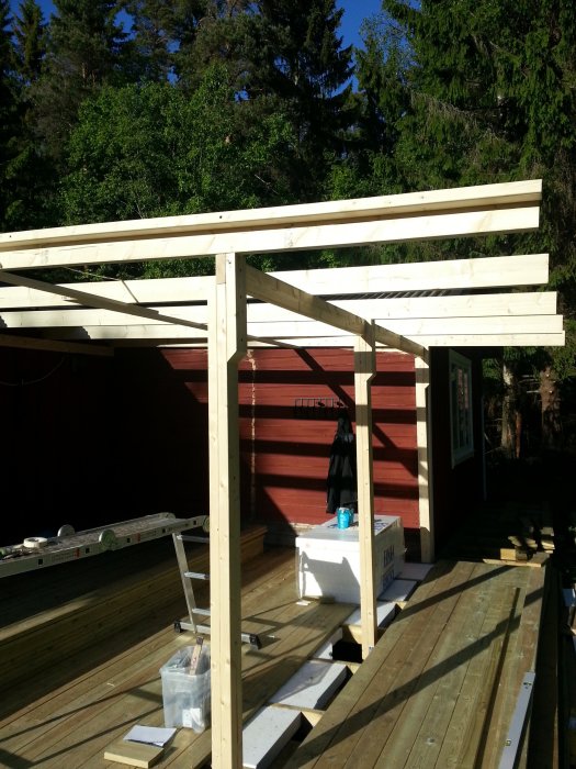 Byggkonstruktion av trä med monterade reglar och bjälkar på en veranda i solljus, omgiven av grönska.