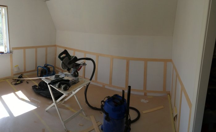 Ett rum under renovering med ommålad vägg och enkel spegelbröstpanel halvfärdig, verktyg utspridda på golvet.
