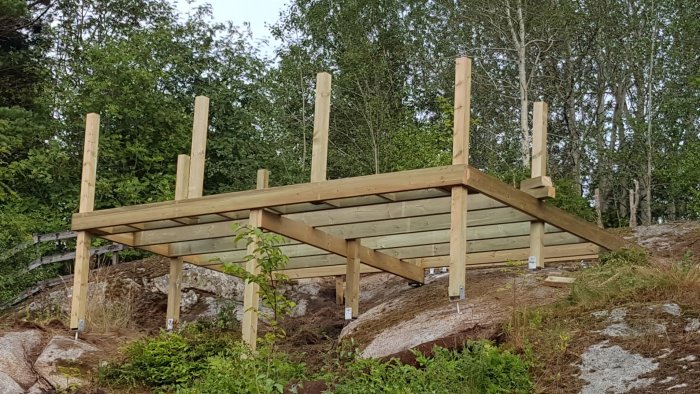 Pågående bygge av en träröstskelettstruktur för en altan på en sluttning med synliga plintar och bärlinor.