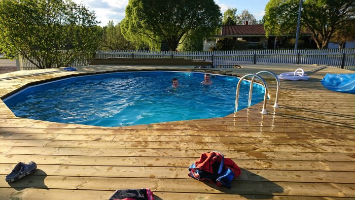 Två personer badar i en utomhuspool med träplattform och badleksaker runt omkring.