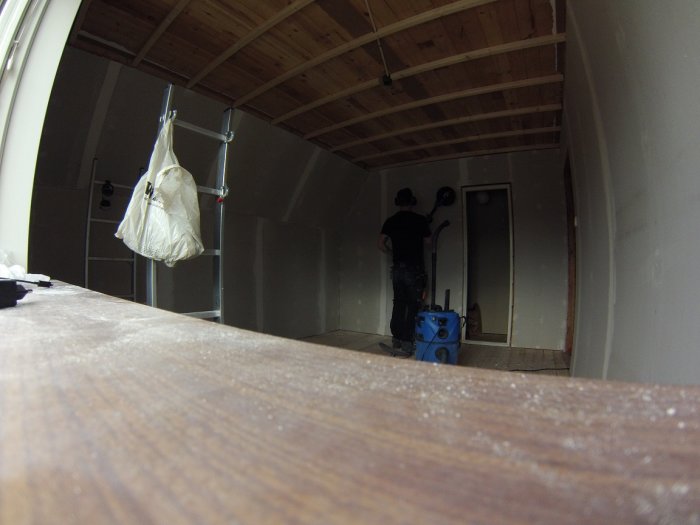 Person använder en väggslipmaskin i ett rum med trägolv och träbjälkar i taket.