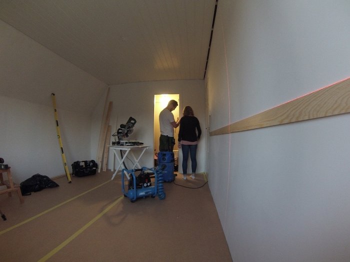 Renoveringsprojekt i rum med två personer, verktyg, och påbörjad spegelbröstpanel på väggen.