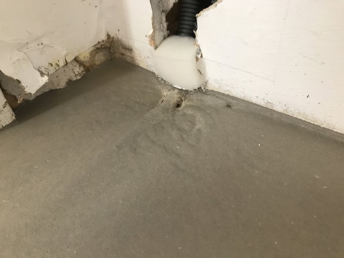 Skadat vägghörn med exponerad isolering och kablar nära ett betonggolv.