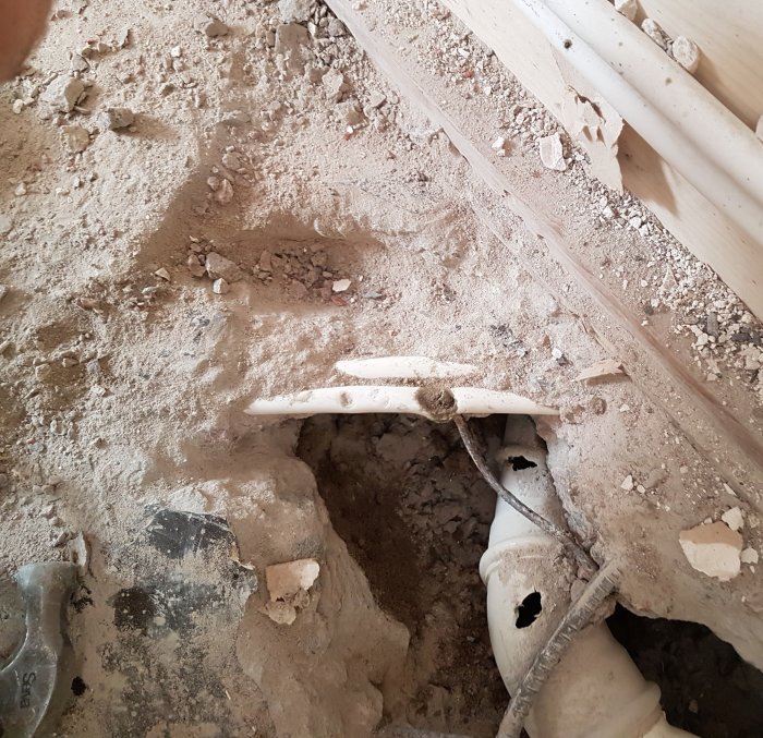 Skadat 12 mm kopparrör synligt i uppbrutet betonggolv bredvid golvbrunn och vit avloppsrör.
