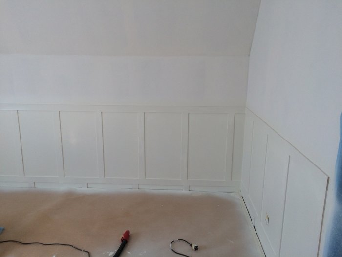 Hörn av ett rum under renovering med nymålat vitt bröstpanel och vägg ovanför täckt med maskeringstejp.