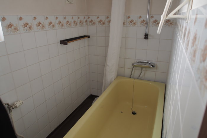 Gammalt badrum med gul badkar, vita kakelväggar med blomstermönster och duschutrustning.