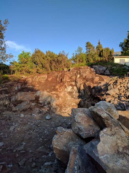 Bergig terräng med stora stenblock och sprängd sten i ett byggprojekt med vegetation i bakgrunden under solnedgången.