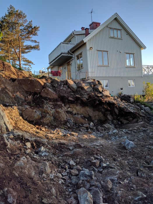 Ett tvåvåningshus med ljust träfasad står nära en brant klippig sluttning med sprängda stenar och grus.