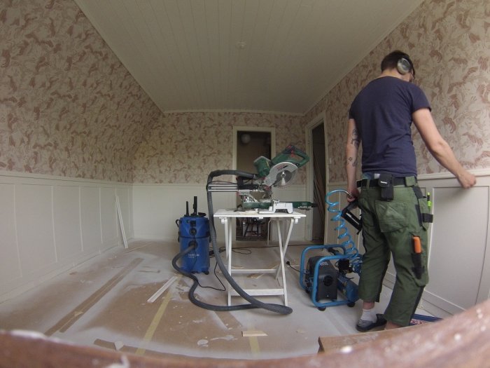 Person tapetserar ett rum med verktyg och utrustning för hemrenovering i förgrunden.
