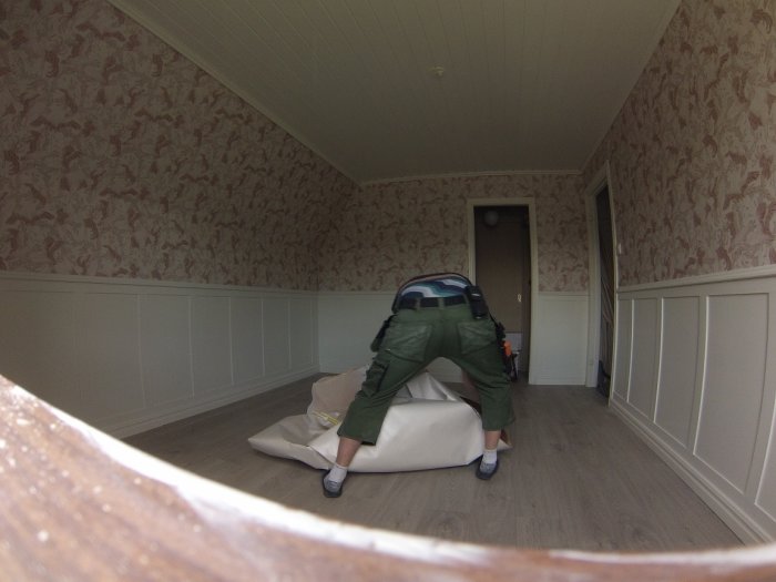 Person tapetserar ett rum med mönstrad tapet och vita panelväggar, taklister syns.