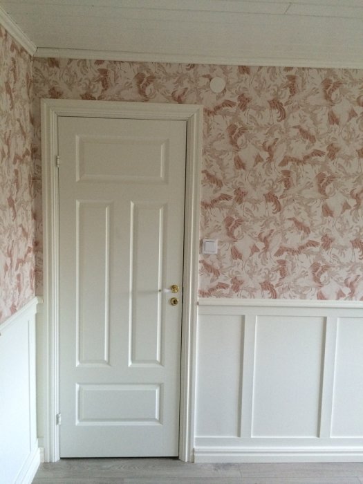 Nyrenoverat rum med vita paneler, tapet med rosa blommönster och en stängd vit dörr, klart för inredning.