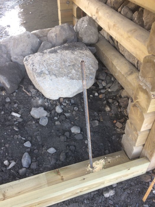 Armeringsjärn står lodrätt i marken bland stenar vid en träkonstruktion vid vattnet.