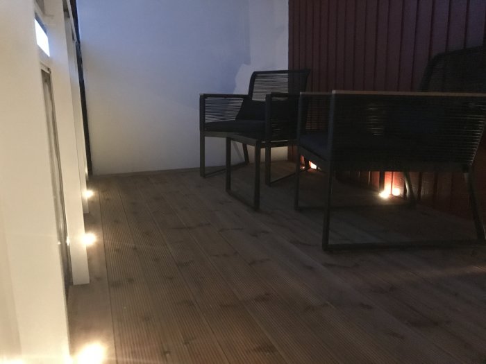 Trallgolv på balkong med reglar under och två svarta trädgårdsstolar, belyst av små lampor.