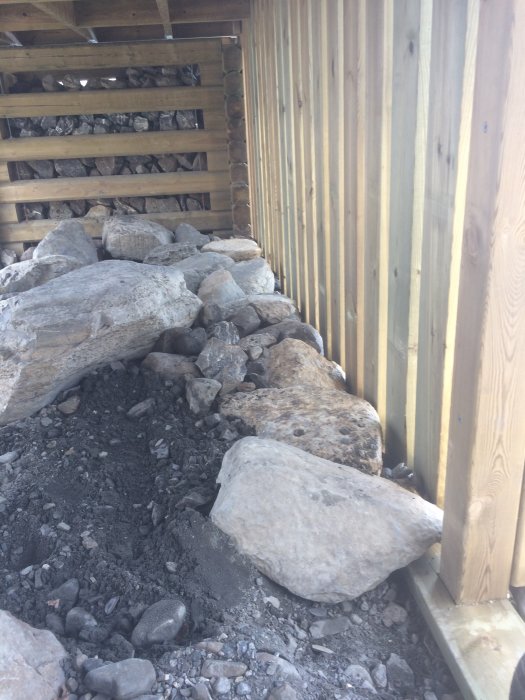 Stödmur med stora stenblock intill trästaket och vedstapel.