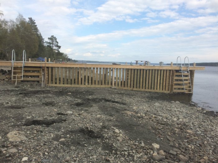 Nybyggd träbrygga vid sjö med räcke, stege och bänk på grusstrand.