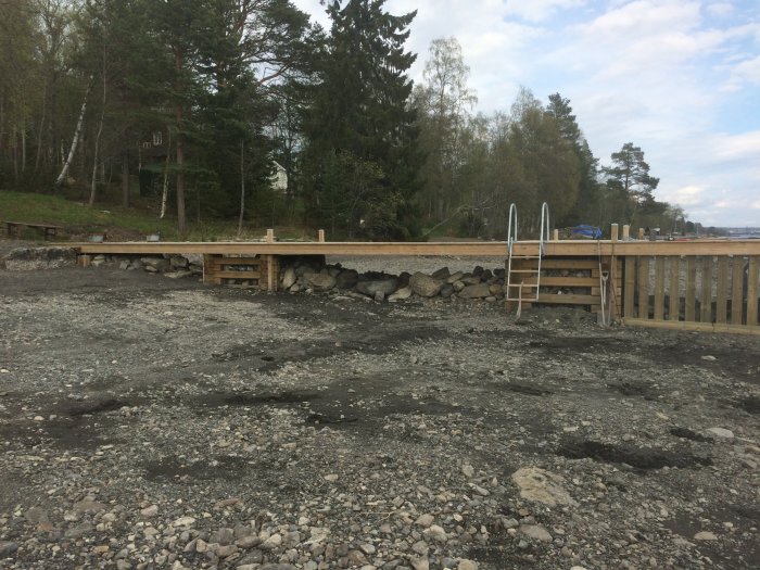 Pågående bygge av träbrygga vid stenig strand med skog i bakgrunden.