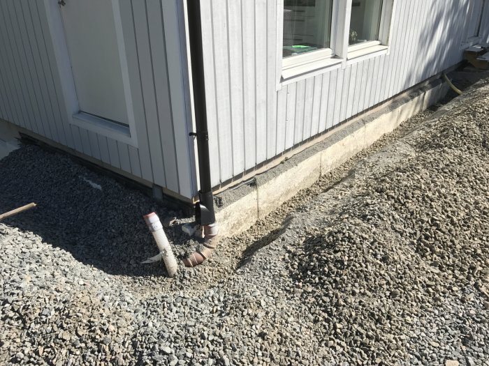 Grävd grop vid husvägg för installation av fiber med synliga fiberrör och grus.