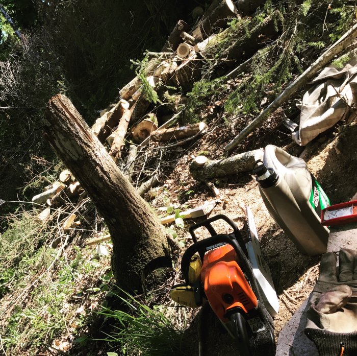 Motorsåg och huggen ved staplad i en skogsmiljö vid husrenovering med handskar och en bränsledunk i förgrunden.