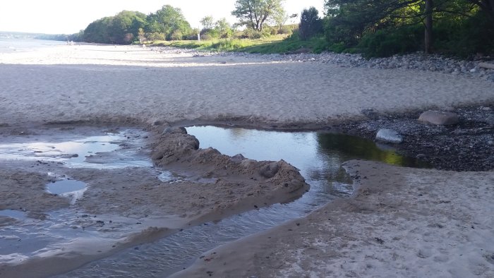 Strand med vattenpöl och sandbank, stenig terräng i bakgrunden och personer i fjärran.