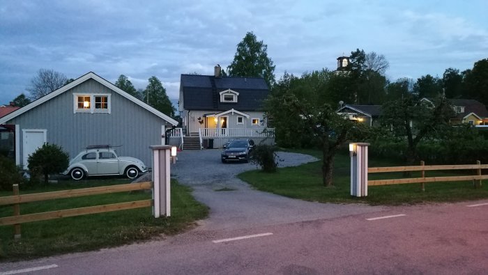 Skymningsbild av uppfart med temporära solcellsdrivna lampor längs staket vid hus med tänt fönster.