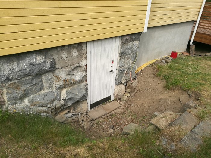 Källarentré med vit dörr, omgiven av ojämn mark och synlig stengrund, med filterduk och gul fiberkabel runtomkring.