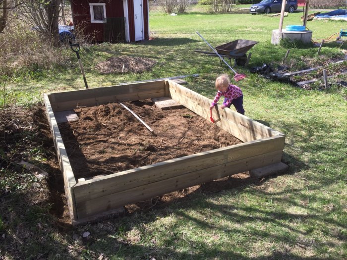 Litet barn leker i ofärdig träramad sandlåda i trädgård, skottkärra och spade i bakgrunden.