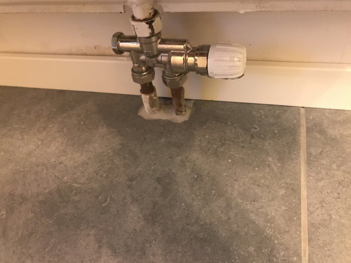 Renoverat golv med rör och termostatventil nära en vägg och vit list.
