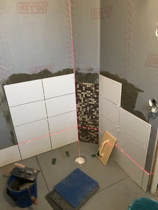 Pågående kakelinstallation i hörn av badrum med delvis satta vita plattor och mosaik samt röd laserlinje.