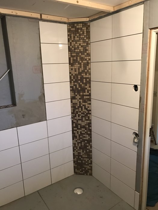 Pågående kakelinstallation i en dusch med vit kakel på väggarna och en accentmosaik i brunt och beige i hörnan.