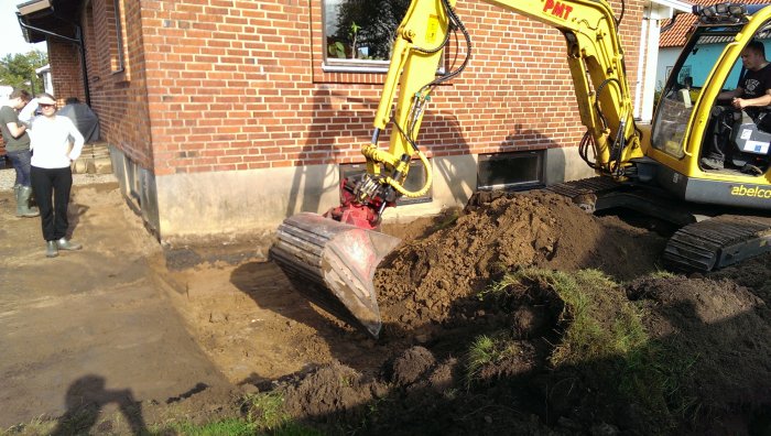 Grävmaskin schaktar för fiberinstallation vid en husgavel på en solig dag.