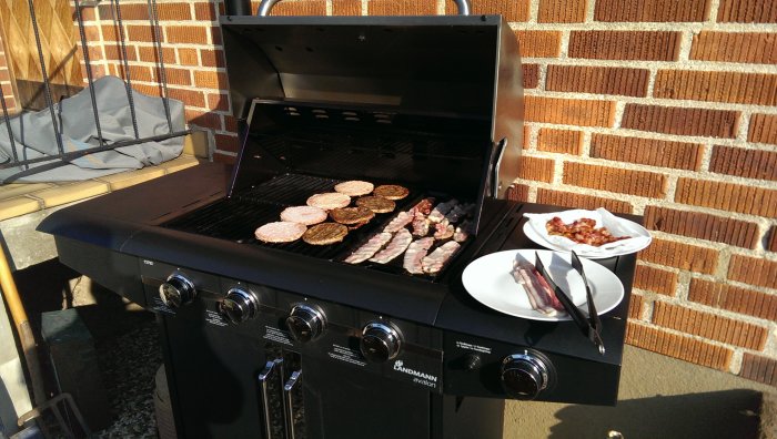 Gasolgrill med hamburgare och bacon under tillagning, uteplatsernas rekreation på en solig dag.