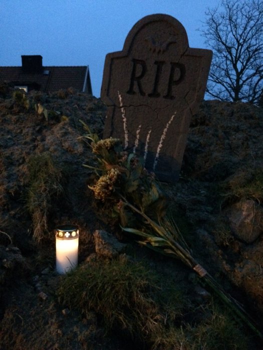 Halloween-dekoration med en RIP-gravsten, vissna blommor och ett tänd ljus i skymningen.