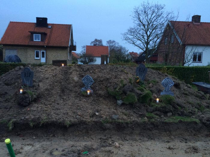 Halloween-dekorationer med gravstenar och ljus på en jordhög framför ett hus i skymningen.