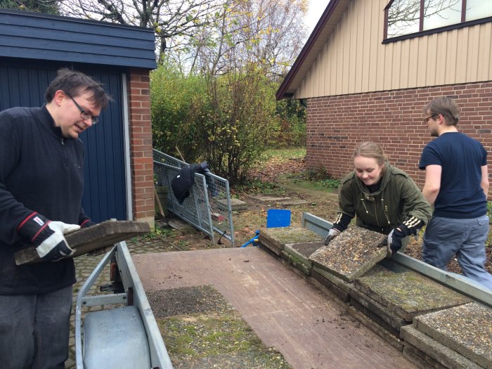 Personer som lastar sjöstensplattor på en släpvagn framför en brun husfasad.