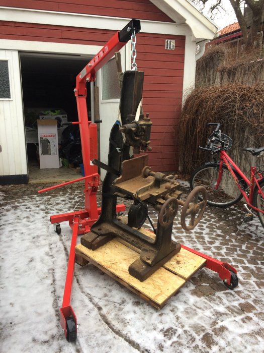 Gammal maskin på en röd lyftvagn framför en öppen garageport med snö på marken och cykel i bakgrunden.