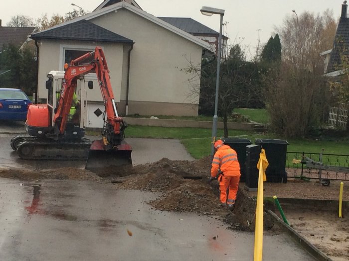 Arbetsfordon gräver för fiberutbyggnad längs en bostadsgata, arbetare synlig, våt väderlek.