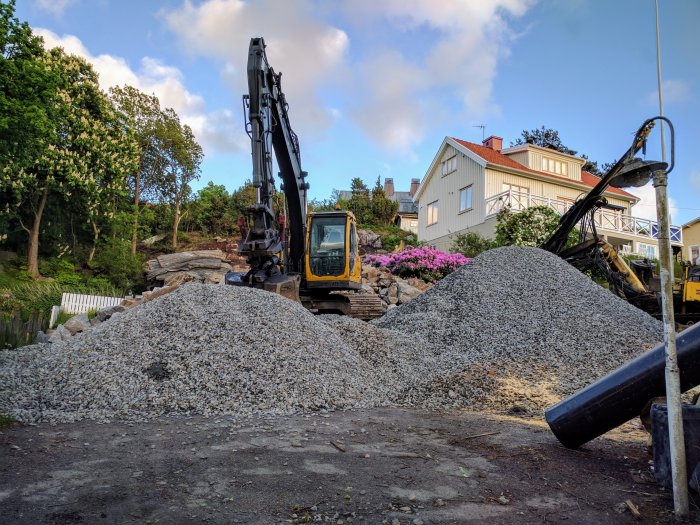 Grushögar framför en grävmaskin på en byggplats med hus och grönska i bakgrunden.