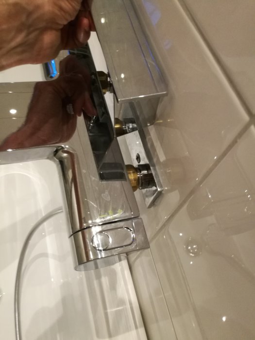 En person rör vid en badrumskran, vit kakelbakgrund, duschslang synlig, bild i vinkel ovanifrån.