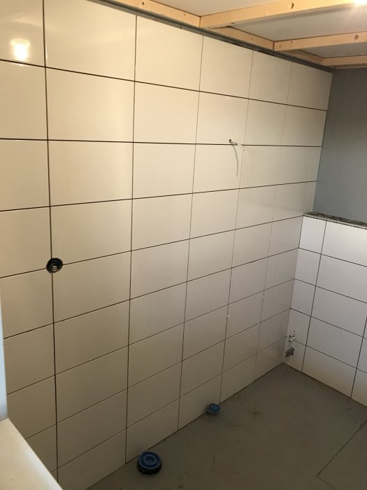 Nyinstallerade vita kakelplattor på vägg i pågående renoveringsprojekt, hörnlist och hylla uppsatta.