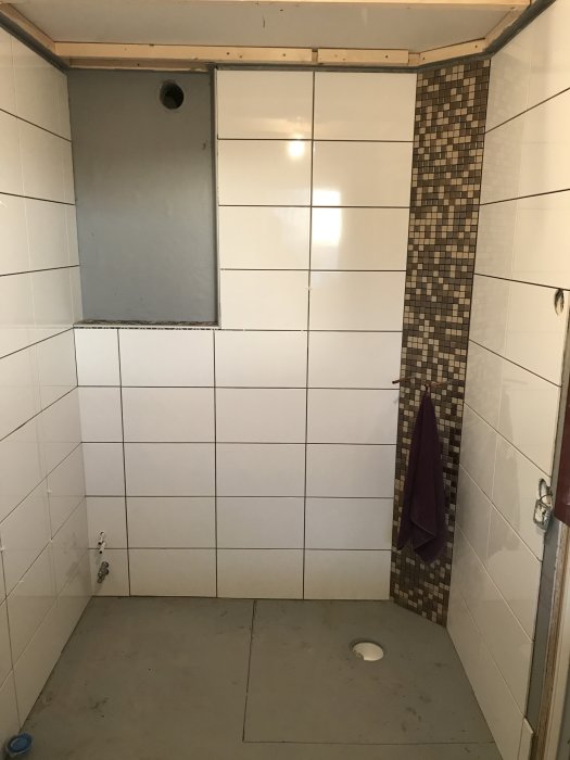 Delvis kaklad duschutrymme med vita väggplattor och mosaik hörnlist, ej färdigställt golv.