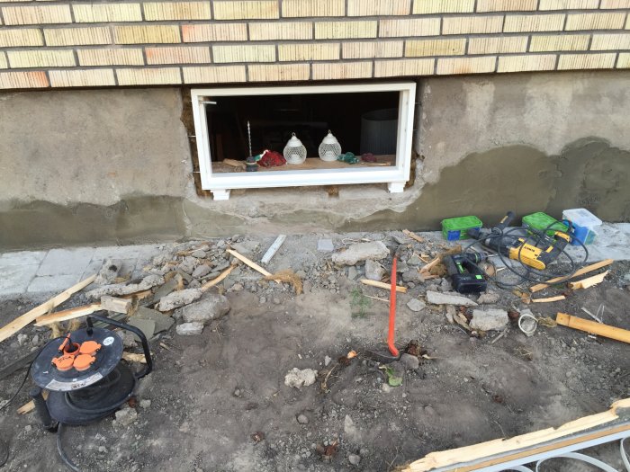 Nyinstallerat fast fönster i putsad fasad med stora drevmånar och byggmaterial och verktyg utspridda på marken framför.