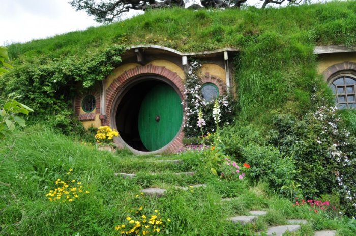 Grönt hobbit-hus med rund dörr och blommor, byggt i en gräsklädd kulle.